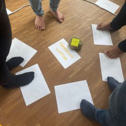 Füße stehen auf Papiersheets im Kreis, in der Mitte liegt ein gelbes Würfel.